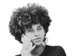 Young person with curly haired styled in an afro. They are wearing a black t-shirt and are holding a smart phone to their ear as if they are talking on the phone.