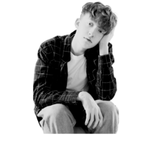 Teen boy with curly hair sits with his hand against the side of his face. He is looking at the camera and is wearing a dark colored plaid shirt.